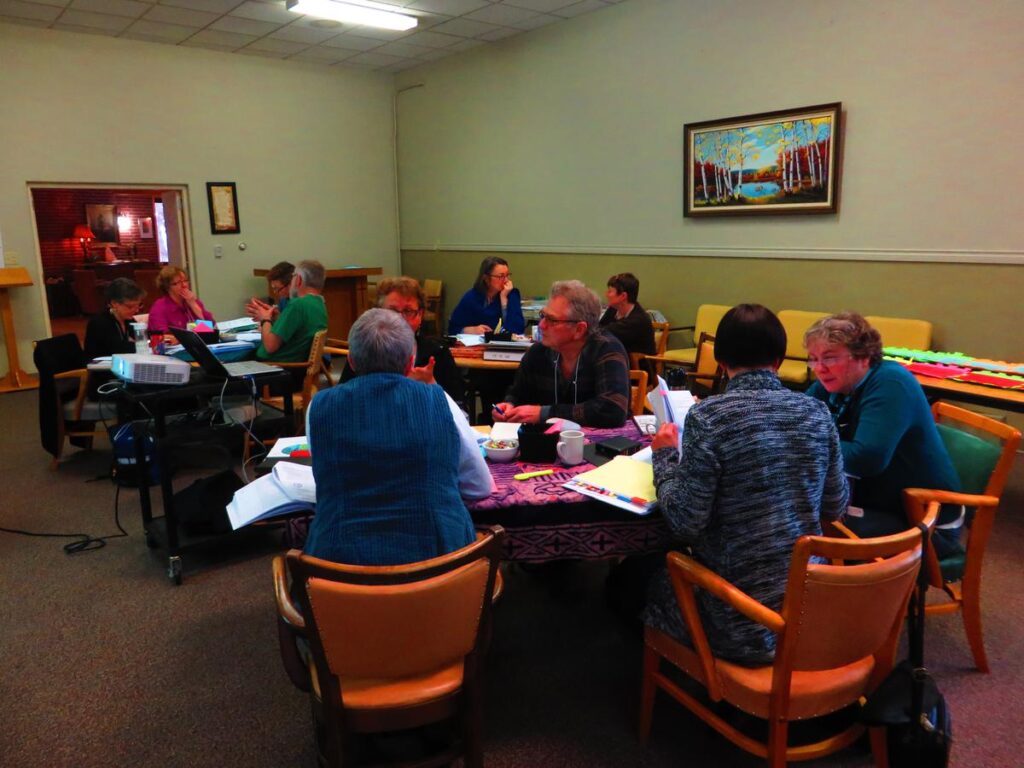 A group of people engaged in a discussion around tables in a community meeting room, with papers and materials scattered on the tables.