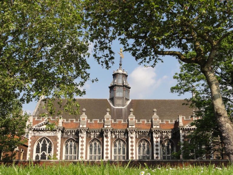Historic building with a clock tower, framed by green trees and a clear blue sky, showcasing architectural details and landscaping.