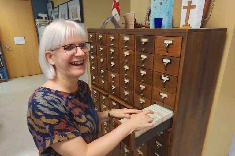 Laurel Parson, a silver-haired woman in her 60s in glasses, smiles at the viewer as she leafs through a drawer of a wooden card catalogue.
