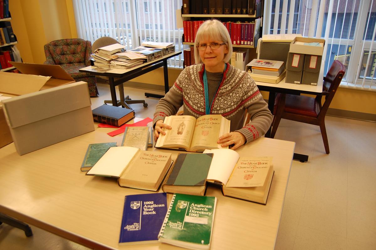 A woman with gray hair, wearing a patterned sweater, sits at a table surrounded by various open books and stacked volumes, in a library setting.