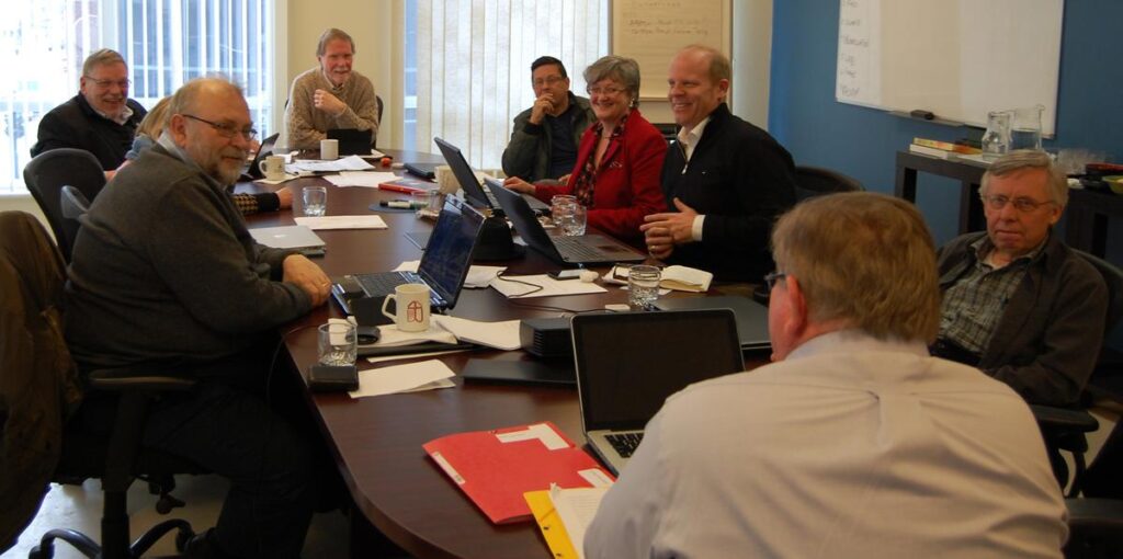 A group of professionals engaged in a meeting around a conference table, discussing various topics with laptops and documents in front of them.