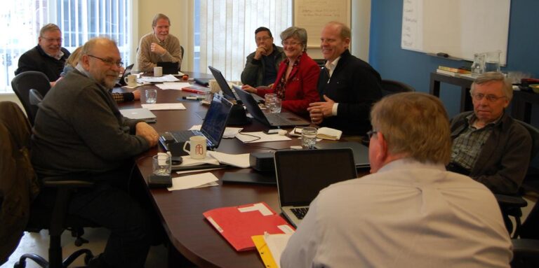 A group of professionals engaged in a meeting around a conference table, discussing various topics with laptops and documents in front of them.
