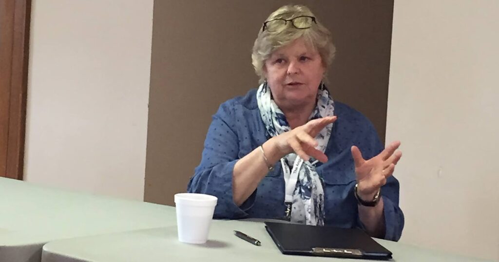A woman with short blonde hair is gesturing while speaking at a table. She is wearing a blue denim jacket and a patterned scarf. A white coffee cup and a notebook are visible on the table.