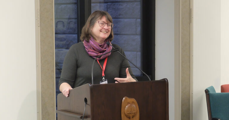 A woman with glasses and a scarf speaks at a podium, engaging the audience with a smile during a presentation or event.