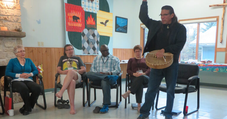 A speaker gestures while presenting to an audience seated in chairs, with colorful banners hanging on the wall in the background. The attendees include three individuals, two men and one woman, engaged in the discussion.