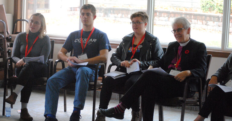 Three individuals seated in a row during a workshop, each wearing a name tag and engaged in discussion. The setting features large windows allowing natural light.