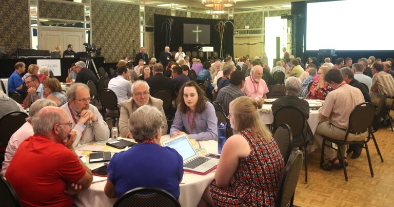 A diverse group of people engaged in discussion around tables during a conference, with a cross displayed in the background.