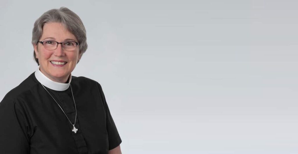 A woman with short gray hair and glasses wears a black shirt, standing in front of a neutral gray background. She appears thoughtful and engaged, conveying a sense of professionalism.