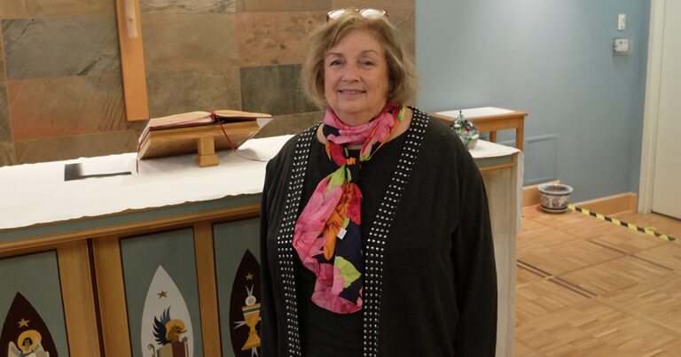 A woman with a colorful scarf stands in front of a wooden altar in a church setting, smiling warmly.