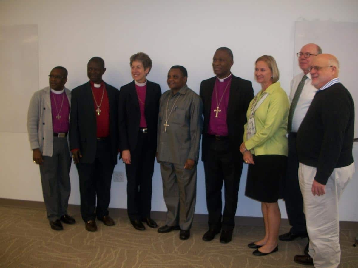 Group photo of six individuals, including religious leaders in clerical attire, posing together in a professional setting. The individuals are smiling and standing side by side, with a neutral background.