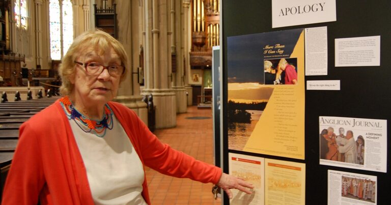 An older woman in a red cardigan points to an information display about a local event in a church setting, with wooden organ pipes visible in the background.