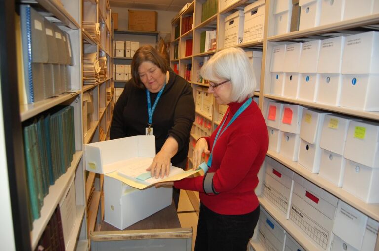 Two women examining documents in a storage room filled with shelves of archival boxes, one holding a folder while the other points at the contents.