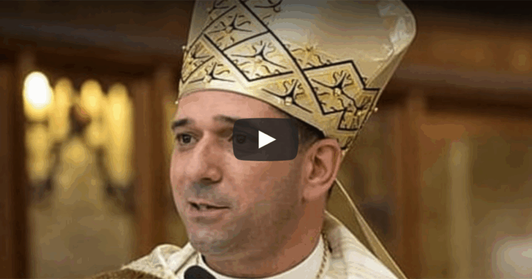 A man wearing a ceremonial bishop's mitre and vestments speaks during a religious ceremony, with a video play button overlay.