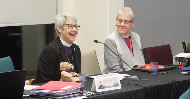 Two women sitting at a conference table, one smiling and holding a stuffed animal, while the other looks on with a thoughtful expression. Papers and a laptop are visible on the table, indicating a meeting or discussion.
