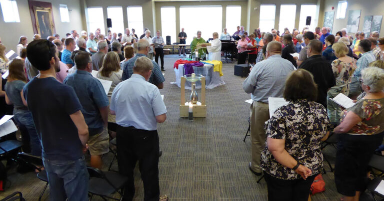 A group of people gathered in a spacious room for a community event, with a table at the front displaying colorful cloths and candles, as attendees engage in a shared experience.