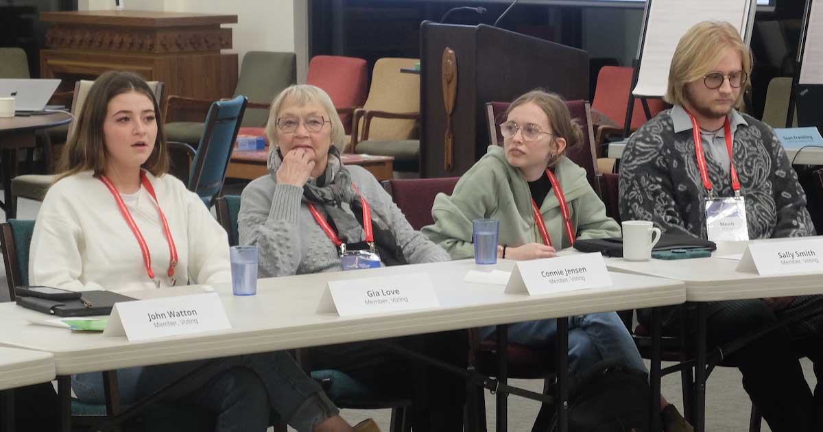 A mid-meeting scene with four people seated at a conference table, each wearing red lanyards. From left to right: a young woman in a white sweater speaking; an older woman in a grey knit sweater resting her hand near her chin; a younger person in glasses and a pale green hoodie looking attentively to the side; and another young adult with long blond hair, glasses, and a patterned sweater, seated quietly with hands folded. Name placards and water cups sit on the table, with stacked chairs and meeting materials visible in the background.