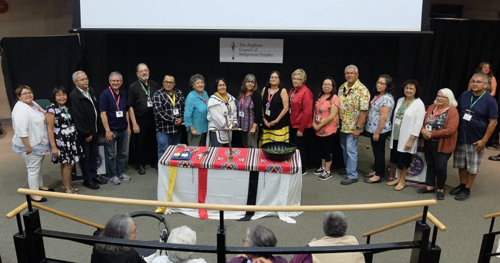 Group of individuals standing together on stage at an event, with a table displaying a colorful cloth and various items. The background features a banner, indicating a formal gathering or ceremony.