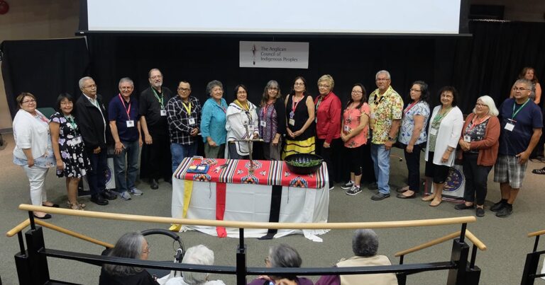Group of individuals standing together on stage at an event, with a table displaying a colorful cloth and various items. The background features a banner, indicating a formal gathering or ceremony.