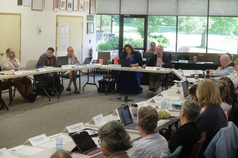 People sit around tables in a conference room, participating in a meeting with laptops and papers; large windows show a green outdoor view.Council of General Synod engages in discussion at their June meeting. Photo: Matthew Puddister