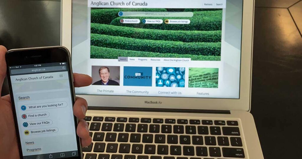 A person holding a smartphone displaying the Anglicans of Canada website on a laptop screen, featuring a green landscape background and various content sections.
