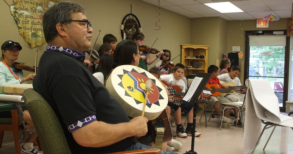 Group of musicians participating in a music class, with a focus on a man playing a drum, surrounded by students playing various instruments. The setting is an indoor classroom, emphasizing cultural music education.