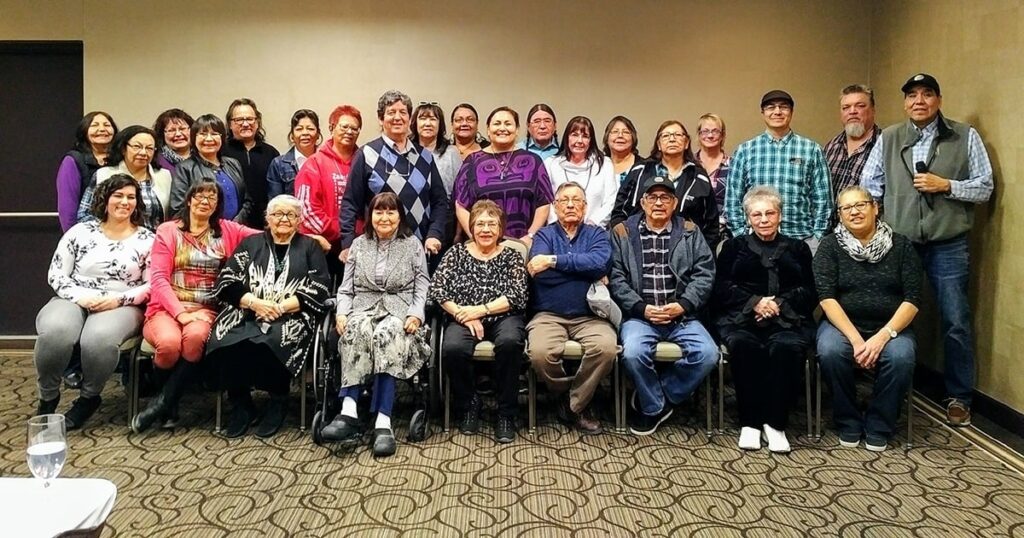 Group photo of a diverse gathering, featuring individuals of various ages and backgrounds, seated and standing together in a conference room setting. The image captures a sense of community and collaboration.