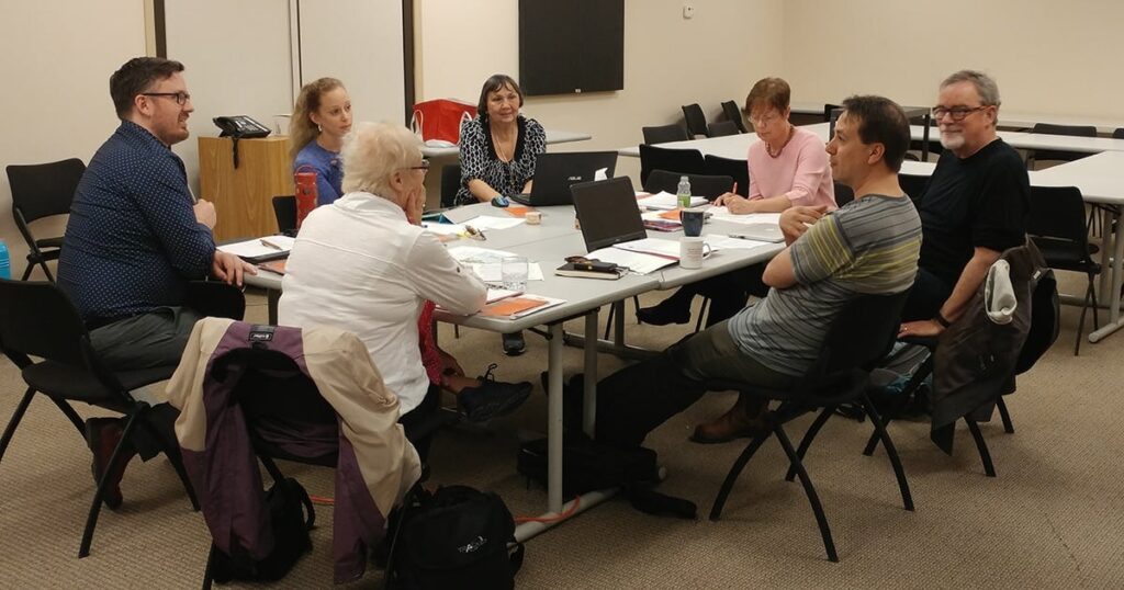 Group of five adults engaged in a collaborative discussion around a table, with laptops and papers visible, in a classroom setting.