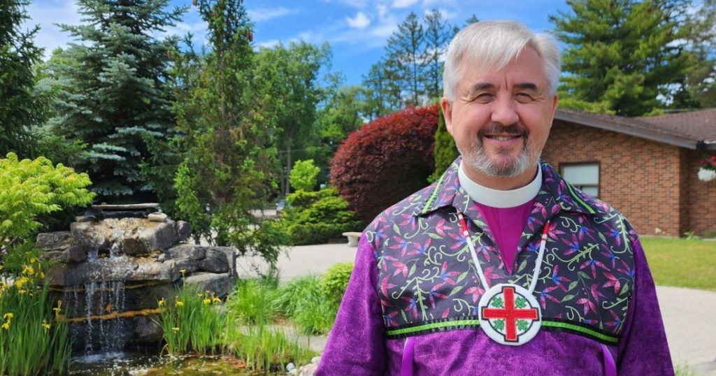 A man with grey hair and a salt-and-pepper goatee in a purple shirt with indigenous canadian patterned fabric stands in front of a pond and artificial waterfall on a sunny day. He is wearing a beaded medallion of the Anglican Church of Canada's Cross logo