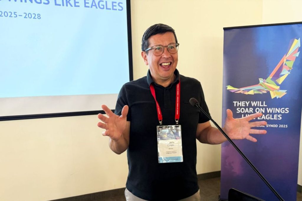 Travis Enright, a middle-aged Indigenous man in a black polo shirt speaks at a podium with a smile on his face and his hands in an open gesture. Behind him, a poster with a multicoloured abstract eagle on it with a legend that reads "They will soar on wings like eagles, General Synod 2025"