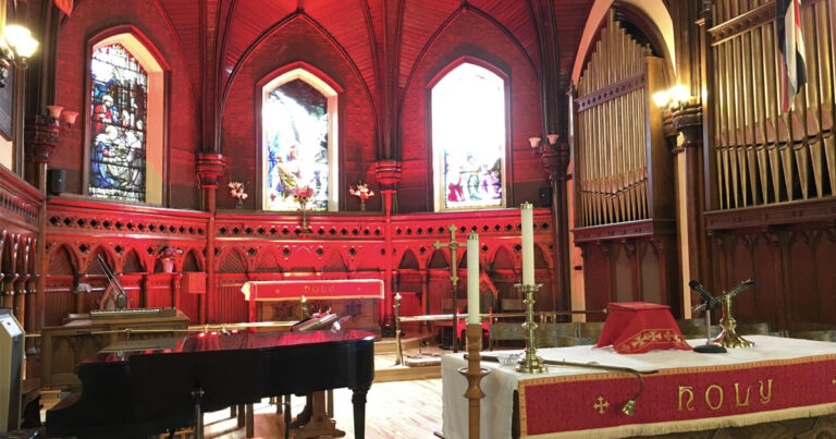 Interior view of a church featuring red lighting, stained glass windows, a grand piano, and decorative candles arranged around the altar.