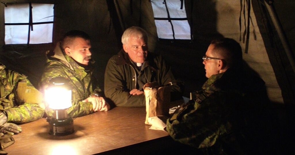 Three men engaged in a serious discussion around a table in a dimly lit tent, wearing military gear. The setting suggests a tactical or strategic meeting.