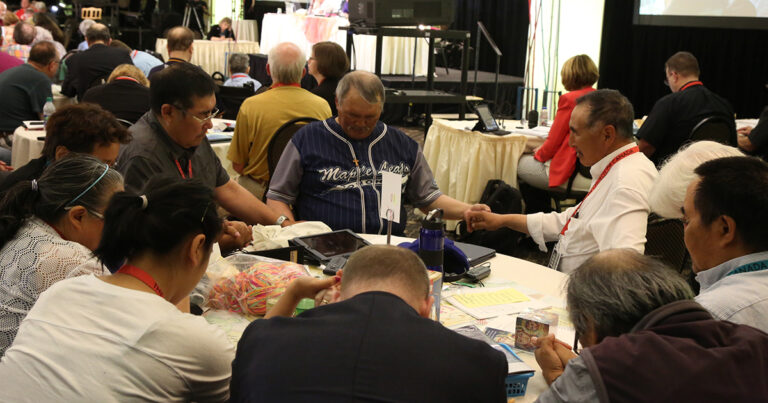 Group of individuals engaged in a collaborative discussion around a table, with some members holding hands in a gesture of unity, while others focus on materials in front of them. The setting appears to be a conference or workshop environment.