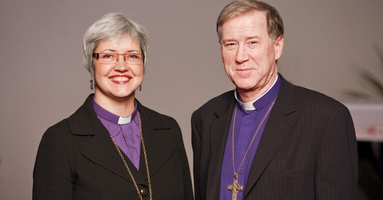 Portrait of two clergy members smiling, one with short gray hair and glasses, wearing a clerical collar and black attire, and the other with light brown hair in a suit and clerical collar, both wearing religious necklaces.