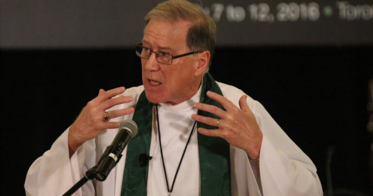 A speaker in a white clerical robe and green stole gestures passionately while addressing an audience at a podium, with a screen displaying text in the background.