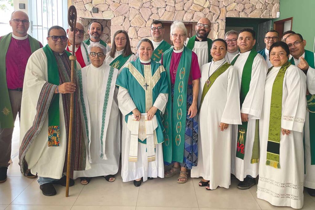 Group photo of clergy standing together indoors, wearing liturgical vestments in green, white, and teal, with one bishop in a magenta shirt and green stole at the centre. The group is smiling and facing the camera.