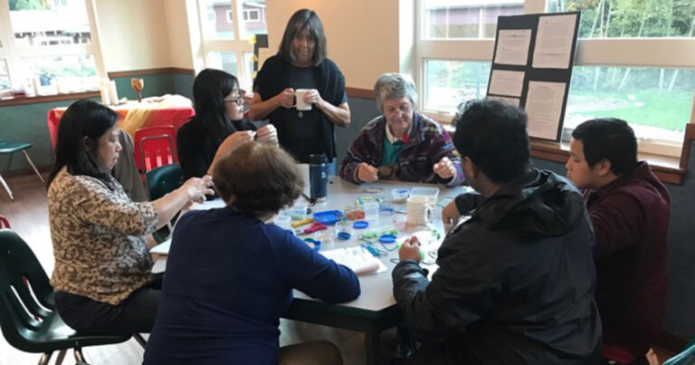 A group of six people engaged in a collaborative activity at a table, with one woman standing and holding a cup while others are focused on colorful materials spread out before them.