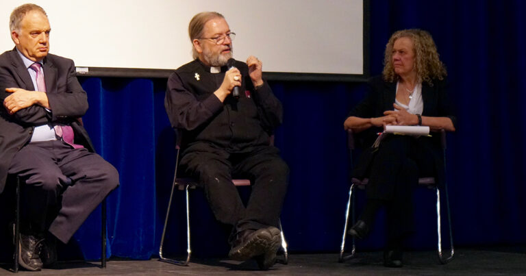 A man in a black vest sits on a stage, engaged in discussion during a panel event, with a woman seated beside him. The background features a projector screen.