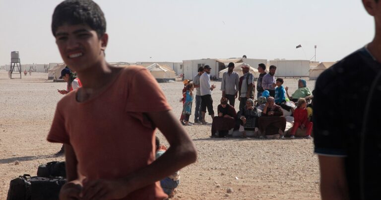 A young boy runs in the foreground of a refugee camp, with a group of people sitting in the background under a clear sky. Tents are visible in the distance, highlighting the camp setting.