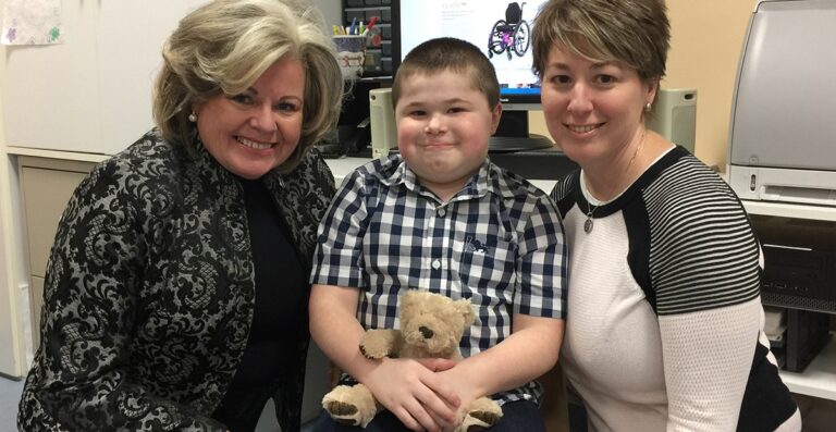 A young boy sitting between two women, holding a teddy bear, smiling for the camera. The background features a computer with an illustration of a wheelchair.