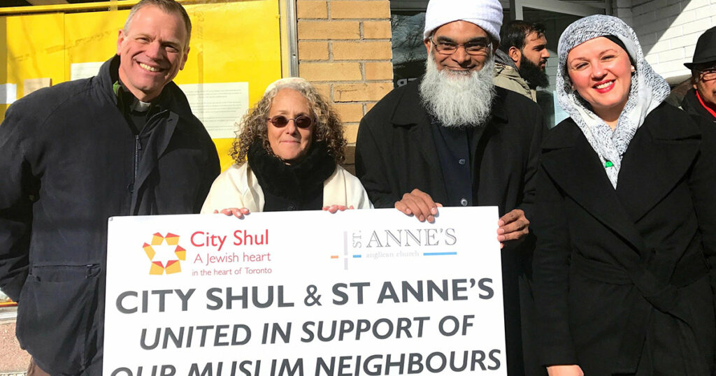 A diverse group of individuals holding a large sign that reads "City Shul & St Anne's United in Support of Muslim Neighbours," demonstrating solidarity and community support.