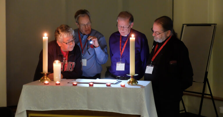 Four individuals gathered around a table illuminated by candles, examining documents under soft light. They appear engaged in discussion, with one person using a flashlight to highlight details. The setting suggests a collaborative or reflective atmosphere.