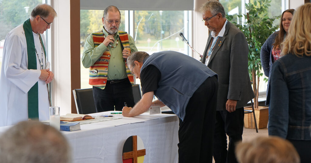 Two men observe as a third man writes on a document at a table adorned with a cross. The setting appears to be a formal or ceremonial event, with natural light coming from large windows in the background.