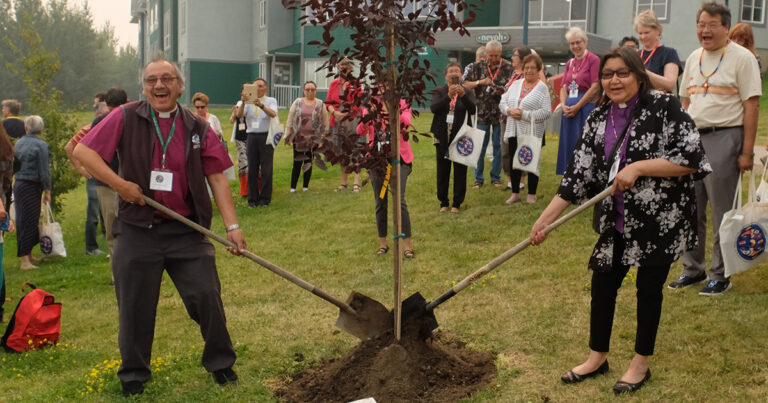 Group of people gathered around as two individuals plant a tree in a grassy area, using shovels to dig around the base.
