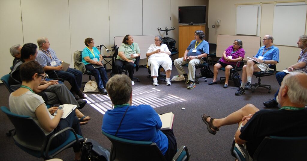 A group of diverse individuals seated in a circle during a discussion in a conference room, with natural light streaming through the blinds.