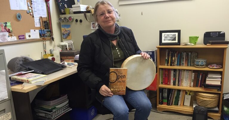 Woman sitting at a desk holding a book titled "Open" in one hand and a drum in the other, with bookshelves in the background.