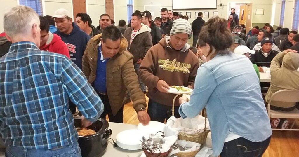 A group of people participating in a community meal event, with individuals serving food from a buffet table. The setting appears lively, with attendees engaging and enjoying the gathering.