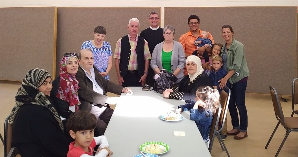A diverse group of people gathered around a table, smiling and engaging with one another in a community setting. The scene includes adults and children, with refreshments on the table, highlighting a social or celebratory event.
