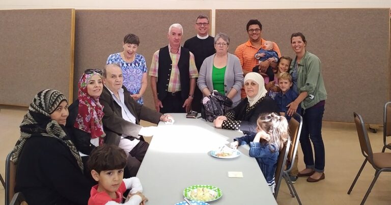 A diverse group of people gathered around a table, smiling and engaging with one another in a community setting. The scene includes adults and children, with refreshments on the table, highlighting a social or celebratory event.