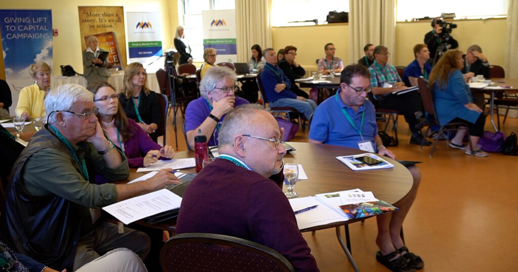 Group of diverse individuals engaged in a workshop setting, seated around tables with notes and materials, actively participating in discussions.