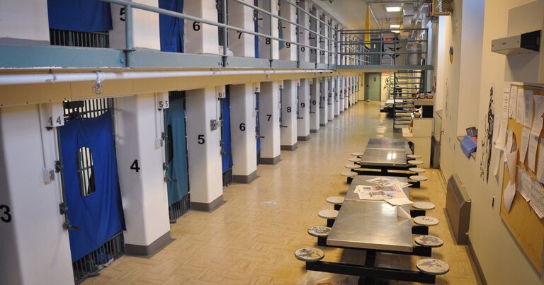 Interior view of a prison corridor featuring multiple cells with closed doors, numbered from 4 to 8, and communal tables along the floor, showcasing the stark environment of correctional facilities.